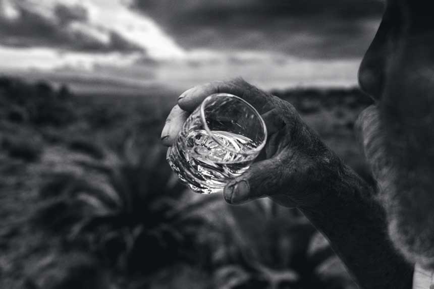 Mano sosteniendo copa de mezcal con agaves al fondo bajo cielo dramático