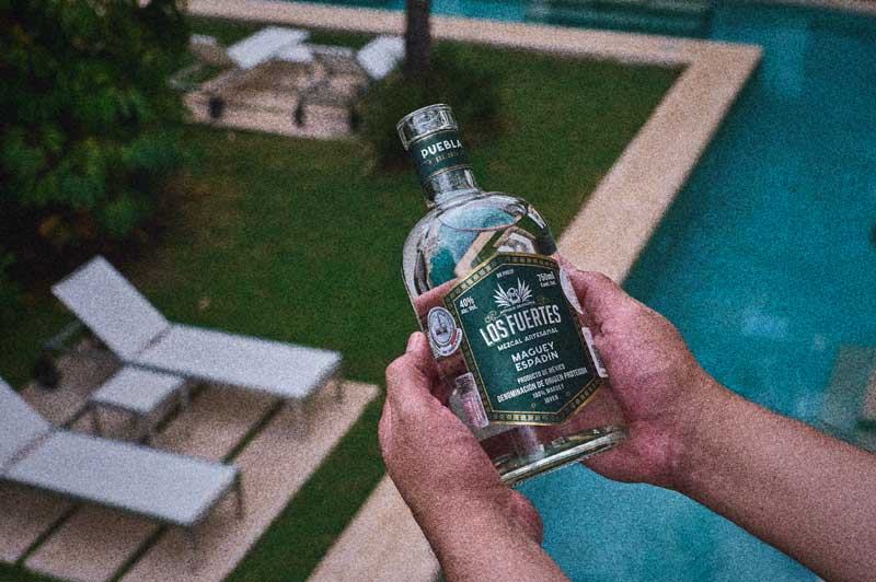 Botella de mezcal Antigua Destilería Los Fuertes de Maguey Espadín sostenida frente a alberca y jardín